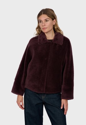 Femme portant une veste en peluche violet foncé avec de larges manches et un col, associée à un jean bleu foncé, debout sur un fond clair.