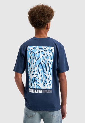 Jongen met krullend haar, gekleed in een marineblauw T-shirt met een wit en lichtblauw abstract koraalontwerp en het woord "BALLIN" op de achterkant.