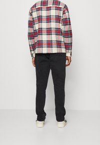 Koszula flanelowa w kratę w kolorze kremowym, czerwonym i niebieskim; zestawiona z czarnymi spodniami. Koszula ma kołnierzyk zapinany na guziki i długie rękawy.