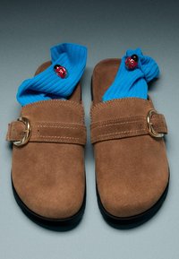 Par brune ruskind slips-on sko med spænder, der indeholder lyseblå ribbestrikkede sokker dekoreret med små røde mariehøne-pins.