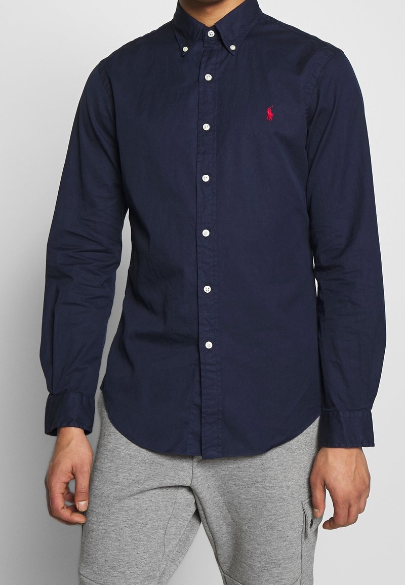 Mörkblå button-up skjorta i bomull, med klassisk krage, långa ärmar och vita knappar. En liten röd logotyp finns på bröstet.