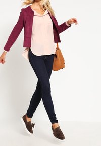 Burgunderfärgad mockajacka över en ljusrosa blus, mörka snäva jeans och bruna slip-on skor med rosetter; brun axelväska.