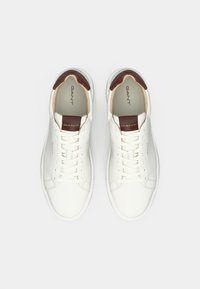 Adidași din piele albă cu o textură netedă, având accente maronii la călcâi și pe limbă, și șireturi albe clasice. Logo-ul GANT se află pe limbă.