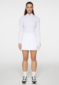 Maglietta bianca a maniche lunghe con colletto alto, abbinata a una gonna bianca svasata. Guanti e scarpe da ginnastica bianco e nere completano il look.