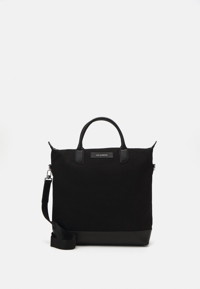 Saco tote preto com corpo em lona, alças de couro e uma alça de ombro destacável. Apresenta uma base plana e um design minimalista.