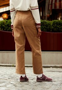 Beige cargo bukser med to baglommer og en løs pasform, parret med mørkerøde sneakers og hvide sokker, sat mod et brostenbelagt underlag.