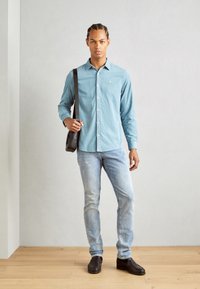 Camisa de botón azul claro, jeans ajustados y zapatos negros. El modelo sostiene un bolso marrón de hombro, de pie sobre un suelo de madera frente a una pared clara.