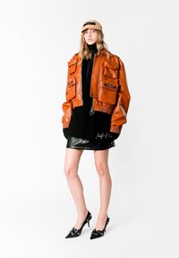 Chaqueta bomber de cuero naranja con múltiples bolsillos, combinada con un vestido de punto negro y tacones de aguja negros brillantes con adornos.
