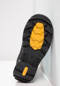 Sola de sapato de borracha preta com sulcos profundos para tração, apresentando um entressola amarelo brilhante com recortes circulares e um detalhe de marca.