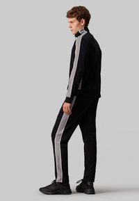 Tuta nera con strisce laterali grigie, colletto con zip e dettagli con logo sulle maniche e sui pantaloni, realizzata in tessuto elasticizzato.