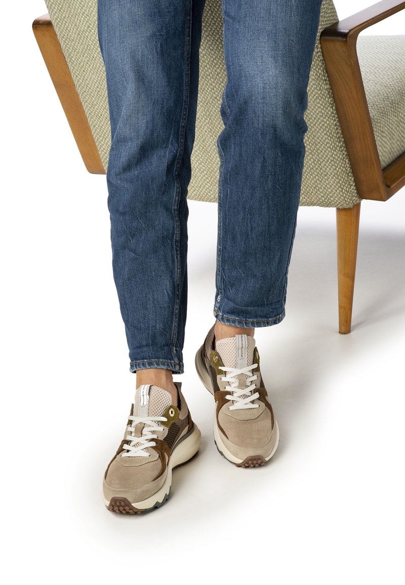 Personne portant un jean bleu et des baskets beige debout à côté d'une chaise en bois avec un rembourrage vert clair sur un sol blanc.