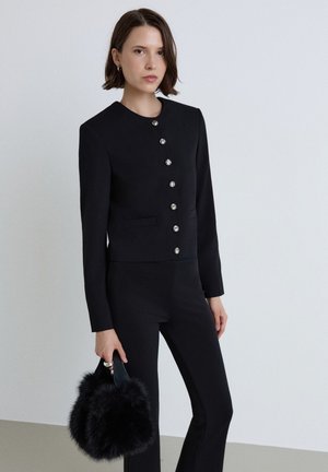 Donna con capelli corti castani che indossa una giacca nera con bottoni e pantaloni, che tiene in mano una piccola borsa nera pelosa su uno sfondo semplice.