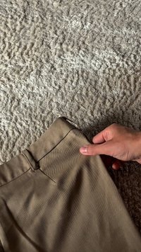 Pantaloni din țesătură bej, cu o textură netedă, având un brâu și o cusătură laterală vizibile. O mână ține articolul pentru a accentua detaliul.