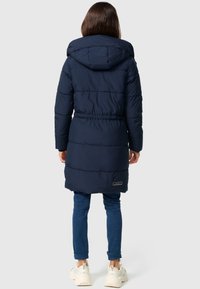 Navy blue padded winter coat with a hood, cinched waist, and side logo patch, paired with blue jeans and light-colored sneakers.