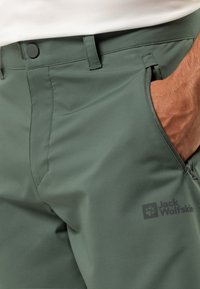 Pantaloni da trekking leggeri e verdi con una texture liscia, dotati di tasche laterali, chiusura con bottone e un logo sulla gamba inferiore.