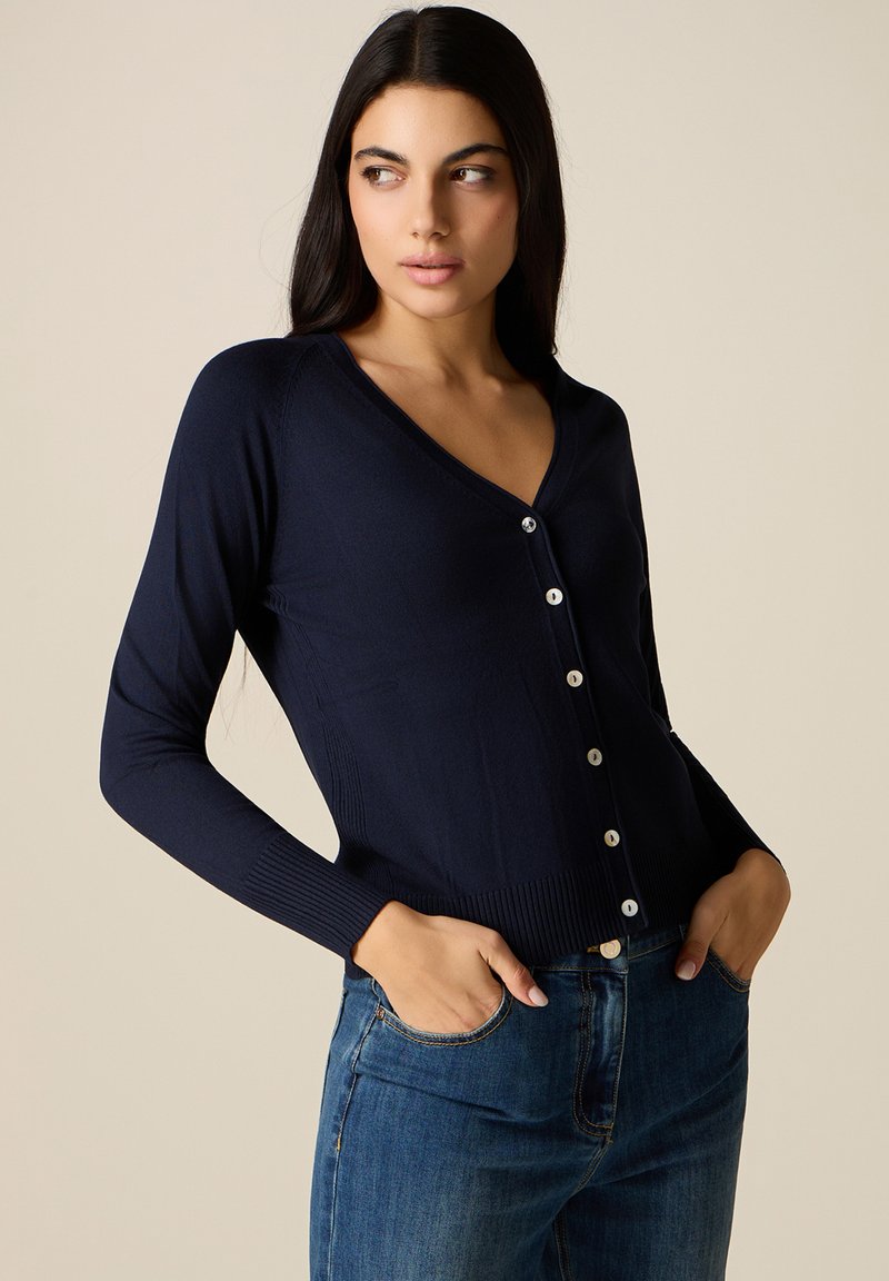 Femme aux longs cheveux bruns portant un cardigan bleu marine boutonné et un jean bleu, les mains dans les poches, regardant sur le côté.