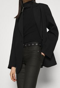 Blazer negro de corte relajado, suéter negro de cuello alto texturizado y pantalones oscuros verdes ajustados. Presenta un cinturón negro con ojales metálicos.