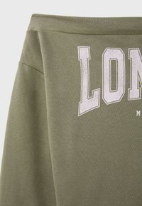 Tissu vert olive avec de grandes lettres blanches usées formant "LON" sur un sweat-shirt ou un t-shirt, montrant la partie supérieure de la poitrine et des épaules.