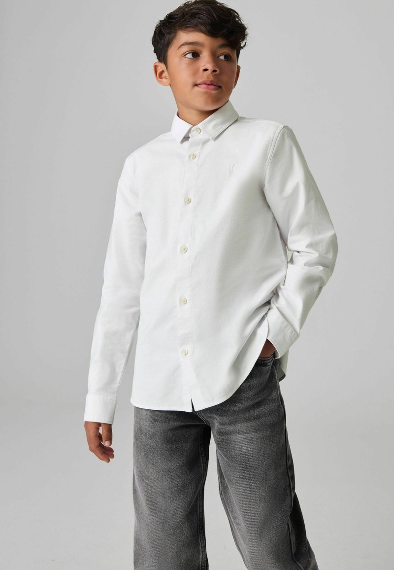 Witte button-up blouse met lange mouwen en een puntige kraag, met een subtiele logo op de borst. Gecombineerd met grijze spijkerbroek.
