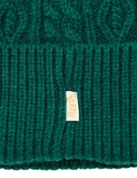 Gebreide groene wolmuts met een ribbelgedeelte en een kabelpatroon. Heeft een klein crèmekleurig label met bruine tekst eraan bevestigd.