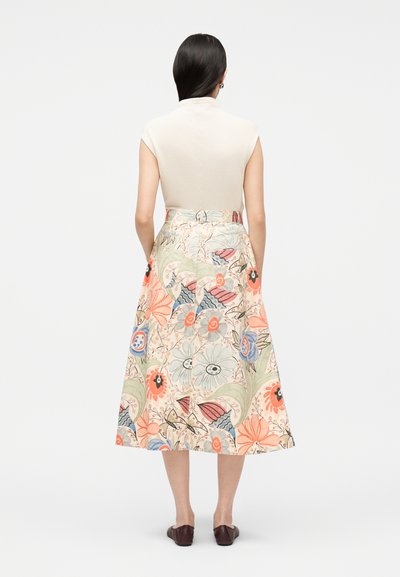 Γυναίκα με σκούρα μαλλιά, φορώντας λευκό αμάνικο τοπ και φούστα μέσης με λουλουδάτο μοτίβο μήκους μέχρι τη μέση της γάμπας, όρθια, με την πλάτη στραμμένη.