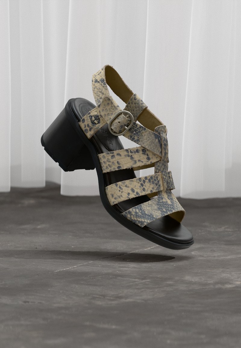 Μπεζ και μαύρο σανδάλι με σχέδιο δέρματος φίδι, με χοντρό μαύρο τακούνι και λουρί αστραγάλου, αιωρούμενο πάνω από γκρι πλακάκια δαπέδου με φόντο λευκή κουρτίνα.