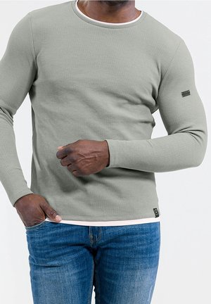 Mann trägt ein hellgraues, strukturiertes Langarmshirt und blaue Jeans, mit der rechten Hand in der Tasche und der linken Hand nahe der Taille.