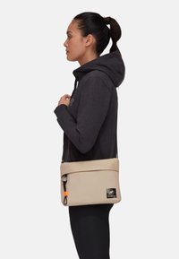 Beige Umhängetasche mit glatter Textur, einem Reißverschluss und einem Logo-Patch. Ausgestattet mit einem verstellbaren Gurt für einen einfacheren Transport.