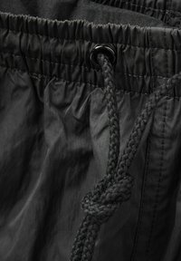 Černý elastický pas s opletenou šňůrkou. Tkanina je hladká s mírně lesklou texturou a disponuje nařasenými záhyby pro flexibilitu.