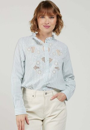 Femme aux cheveux courts et ondulés portant un chemisier bleu clair à rayures avec une broderie florale ajourée et un pantalon couleur crème, la main dans la poche.