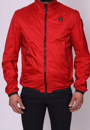 Mężczyzna ubrany w czerwoną kurtkę na zamek z czarnym zamkiem błyskawicznym i elastycznymi mankietami, w połączeniu z czarnymi spodniami, stojący na jednolitym tle.