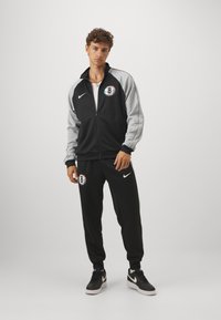 Μαύρο και γκρι προπόνητο πουλόβερ από λεία ύφανση, με φερμουάρ στη ζακέτα, παντελόνι σε αθλητική γραμμή και κυκλικό λογότυπο στο στήθος.