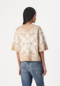 Beige t-skjorte med korte ermer og rund hals, utsmykket med hvite intrikate paisleymønstre, kombinert med blå jeans.