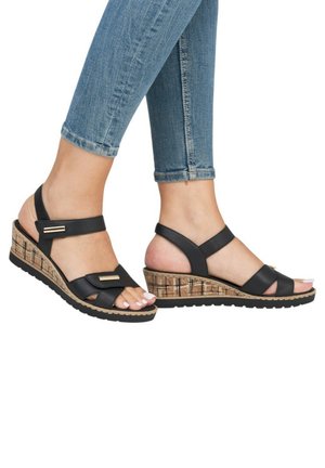 Pieds portant des sandales compensées noires avec des semelles texturées en liège, associées à un jean skinny bleu retroussé sur fond blanc.