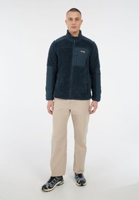 Man staat gekleed in een marineblauwe fleecejack met ritszakken, beige broek en grijze wandelschoenen tegen een effen witte achtergrond.