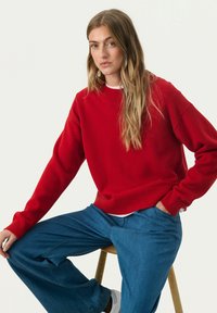 Red knitted sweater with a round neckline and ribbed cuffs, paired with blue wide-leg jeans. Model seated on a stool.