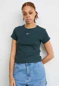 Tamsiai žaliaribota medvilninė T-marškiniai su trumpomis rankovėmis ir apvalia iškirpte. Centre ant krūtinės yra balta Nike logotipas.