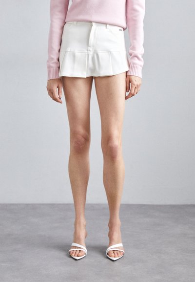 Hvid plisseret mini-nederdel med frontlommer og en glat tekstur, parret med hvide strophæle og en lys pink sweater.