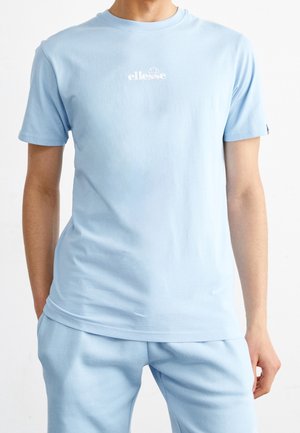 Man wearing a light blue ellesse t-shirt and matching pants, standing with hands partially in pockets against a white background.