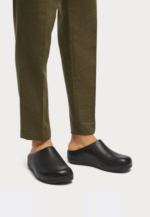Black clogs with a smooth leather finish, rounded shape, and open back, worn with olive green tailored pants.