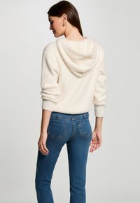 Suéter con capucha de color crema y textura suave, de mangas enrolladas, combinado con jeans ajustados de color azul con bolsillos traseros. Textura suave y diseño casual.