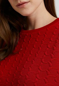 Röd texturerad tröja med ett upphöjt geometriskt mönster och ribbad halsringning. Materialet verkar mjukt med intrikata detaljer genom hela plagget.