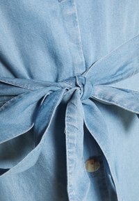 Λεπτομέρεια από ανοιχτό μπλε τζιν πουκάμισο με δεμένο μέσο, κουμπί κλεισίματος και μαλακό, υφασμάτινο υλικό. Κοντινό σε κόμπο και πτυχές του υφάσματος.