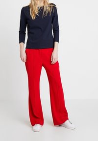 Námořnický top s dlouhým rukávem v kombinaci s jasně červenými širokými kalhotami, které mají jemné pruhy na přední straně, a bílými teniskami. Hladký povrch látky.