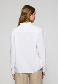 Camicia bianca con colletto, maniche lunghe, realizzata in tessuto leggero, con una trama sottile e orlo arrotondato. Vista posteriore che mostra linee pulite.