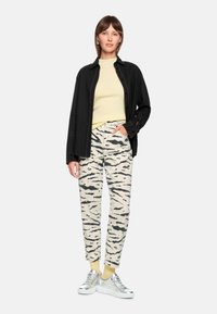 Schwarze knöpfbare Jacke, hellgelber, gerippter Pullover, cremefarbene Hose mit schwarzen Tigerstreifen und silberne Sneakers mit gelben Socken.