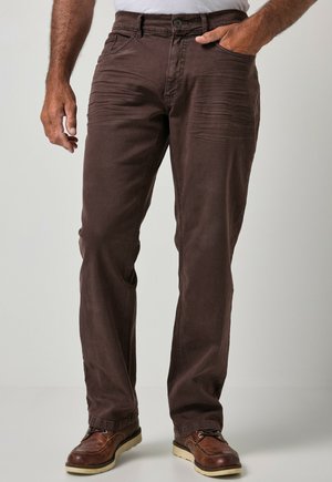 Hombre con pantalones marrones de corte recto con bolsillos delanteros, combinados con zapatos marrones con cordones sobre un fondo liso.