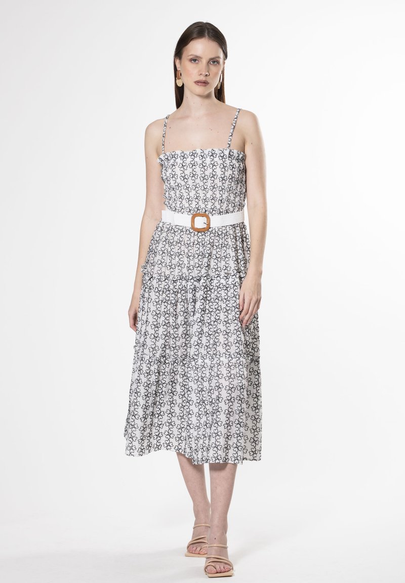 Vestido blanco con estampado floral negro, escote con volantes, tirantes finos y falda con capas. Presenta un cinturón blanco con una hebilla cuadrada marrón.