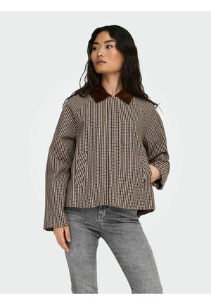 Femme portant une veste à carreaux marron avec un col marron foncé et un jean gris, debout avec une main dans la poche sur un fond blanc.