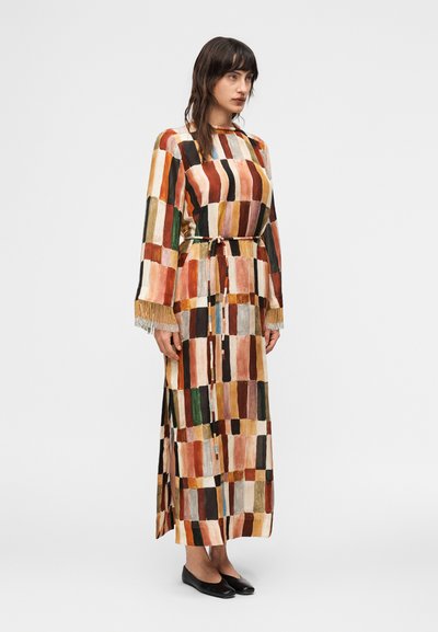 Γυναίκα που στέκεται, φορώντας ένα μακρύ, πολύχρωμο φόρεμα παζλ με μανίκια με φράντζες και λεπτή ζώνη, φορώντας μαύρα επίπεδα παπούτσια σε απλό φόντο.